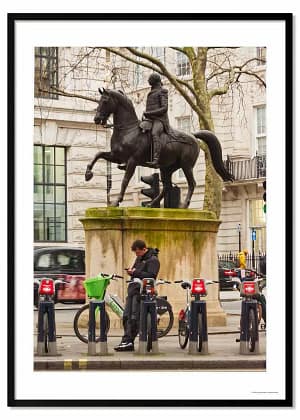 see through walls framed photography poster print called 'Saving Kept Sports', featuring an lone figure leaning on a London Lime Bike as he looks at his phone, while above him is a statue of King George astride a horse.