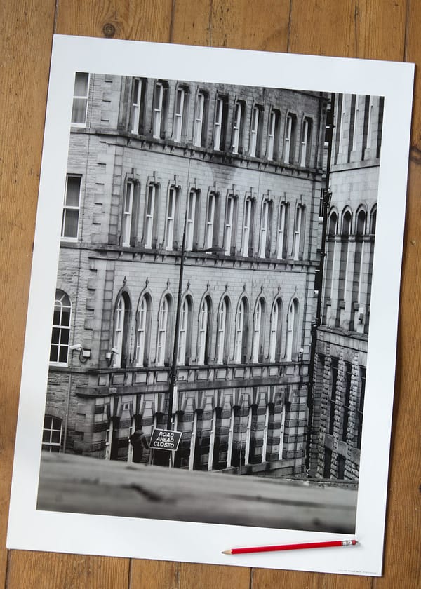 Road Ahead Closed Poster featuring towering historic city buildngs converging to create a narrow perspectice with a roiad sign indicating the Road is Closed.
