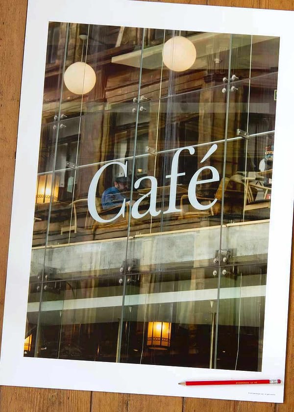 A 70x50cm architectural photography print titled 'Wiring Jolly Beard' featuring a modernist café exterior. Reflections of older urban buildings overlay a view of the interior's transitioning floor levels, industrial lighting, and a lone figure seated behind a large white letter 'C' from 'Cafe' signage.
