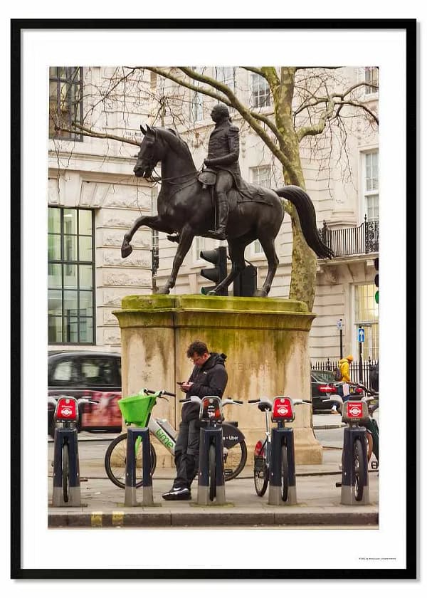 see through walls framed photography poster print called 'Saving Kept Sports', featuring an lone figure leaning on a London Lime Bike as he looks at his phone, while above him is a statue of King George astride a horse.