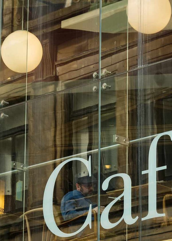 The photograph is a complex architectural study of transparency and reflection. In the foreground, the bold, sans-serif white typography of a café sign is fixed to a large glass pane. Behind the glass, a person sits in profile, framed perfectly by the curve of the letter 'C'. The interior reveals a contemporary industrial space with visible structural beams and warm, spherical pendant lights. Overlaid across this scene is a sharp reflection of the street behind the photographer, showing the textured brickwork and windows of a heritage building, creating a visual bridge between old and new urban design.