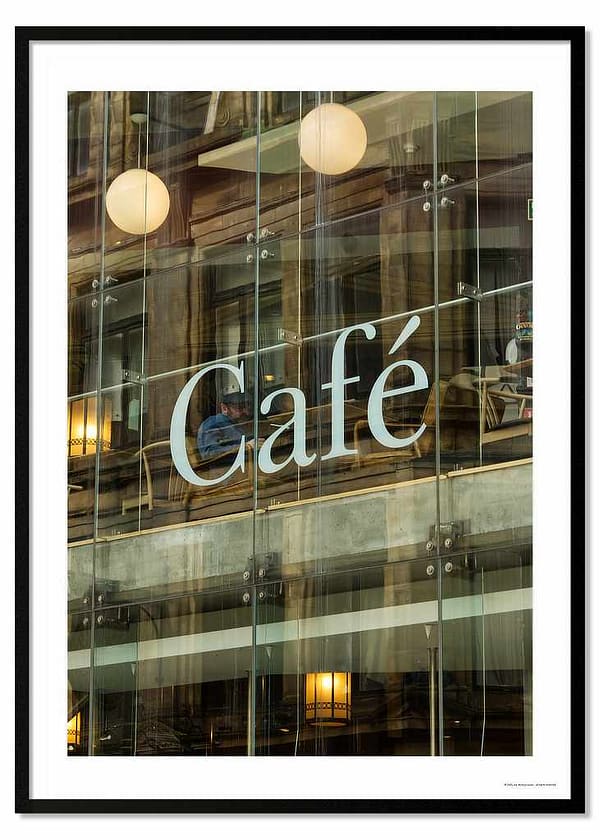 A 70x50cm architectural photography print titled 'Wiring Jolly Beard' featuring a modernist café exterior. Reflections of older urban buildings overlay a view of the interior's transitioning floor levels, industrial lighting, and a lone figure seated behind a large white letter 'C' from 'Cafe' signage.
