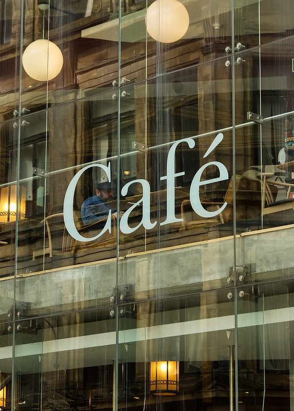 A 70x50cm architectural photography print titled 'Wiring Jolly Beard' featuring a modernist café exterior. Reflections of older urban buildings overlay a view of the interior's transitioning floor levels, industrial lighting, and a lone figure seated behind a large white letter 'C' from 'Cafe' signage.