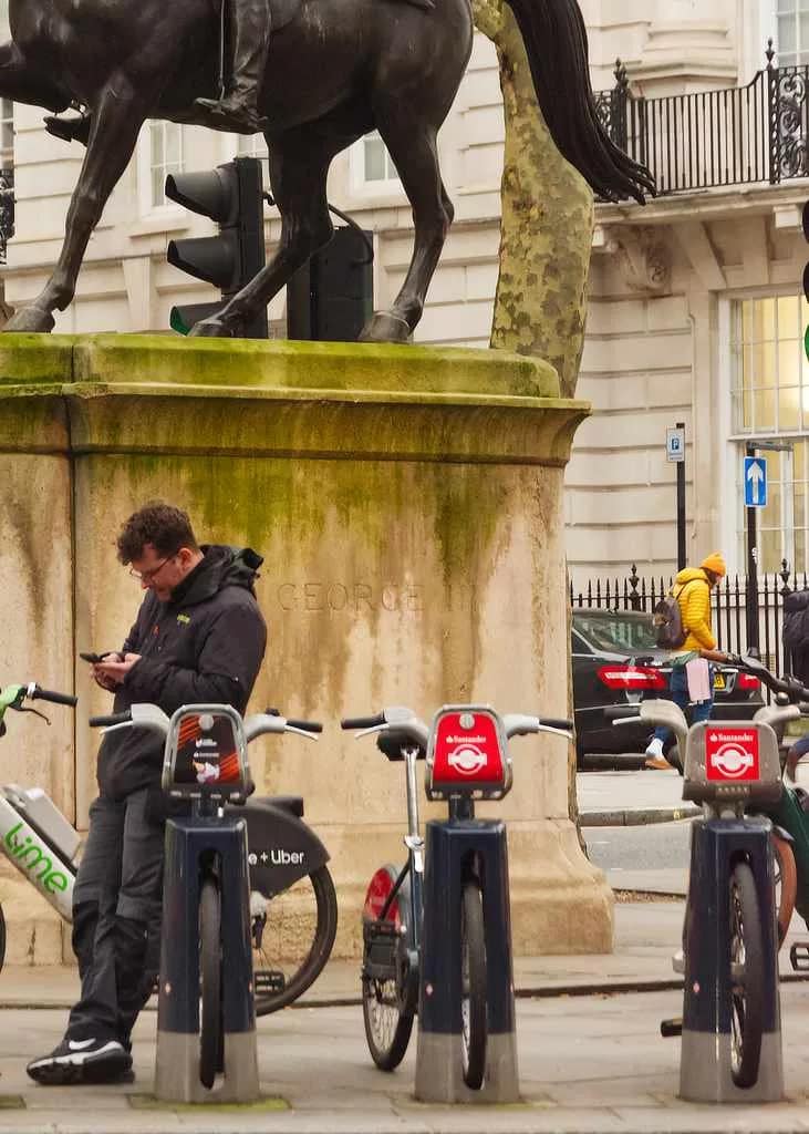 see through walls close up of a photograph called 'Saving Kept Sports', featuring an lone figure leaning on a London Lime Bike as he looks at his phone, while above him is a statue of King George astride a horse.